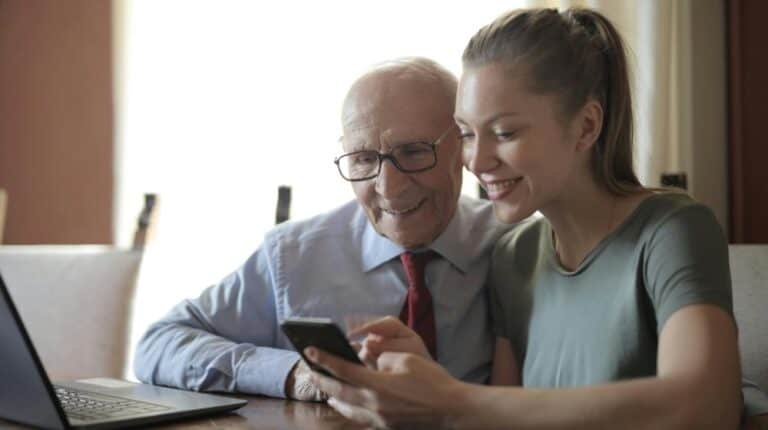 Young positive lady showing photos on smartphone to senior man while sitting at laptop | teach computer skills to seniors px featured image